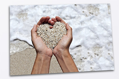 Postkarte auf der man eine Hand sieht mit Sand, geformt zu einem Herz über dem Meer