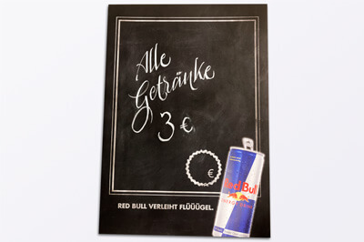 Ein Plakat mit schwarzer Tafelfarbenveredelung, unten rechts auf dem Plakat eine Dose Red Bull und auf dem Plakat steht "Alle Getränke 3€".